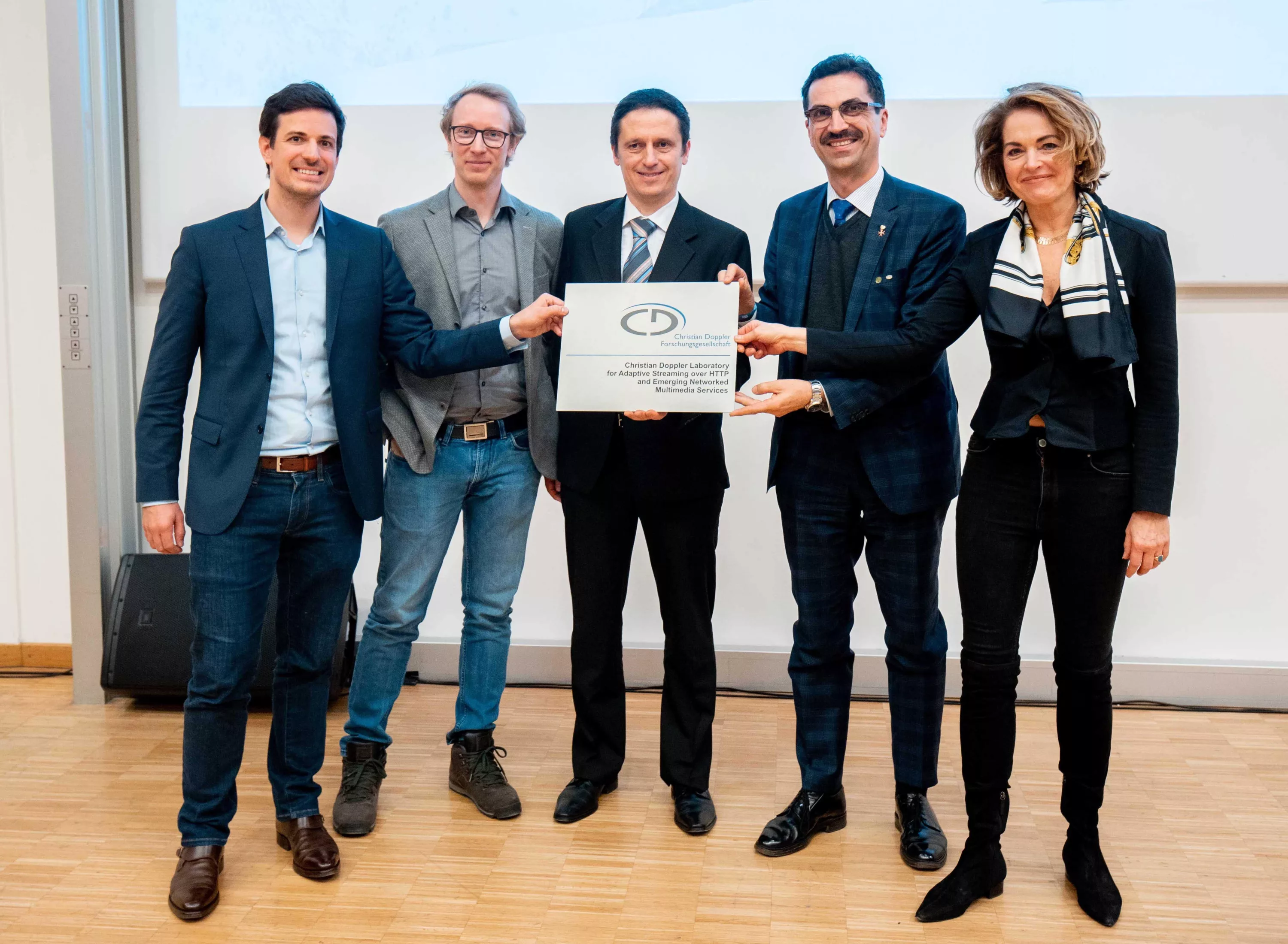 Bitmovin co-founders Stefan Lederer, Christopher Mueller, and Christian Timmerer celebrating the opening of the Christian Doppler ATHENA Laboratory for video research, holding a sign/plaque for the Lab's entrance together with Martin Gerzabek and Ulrike Unterer from the Christian Doppler Research Association.